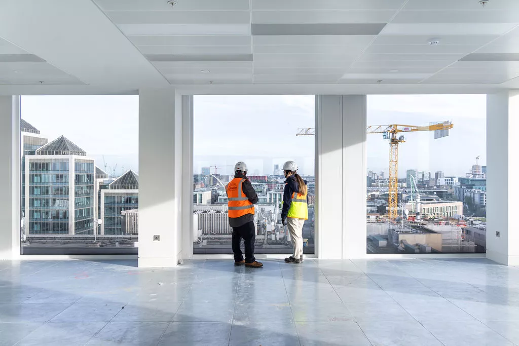 A mans and women both wearing white hard hats and Ashview Consultants high visability jackets on the 10th floor, standing in centre of three large floor to ceiling window in a new office building. The view from the window is of office building beside the river liffery in Dublin Ireand. There is a large yellow construction crain in the right window view.
